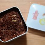 Boîte métallique contenant du rooibos, couvercle décoré, posé sur bois